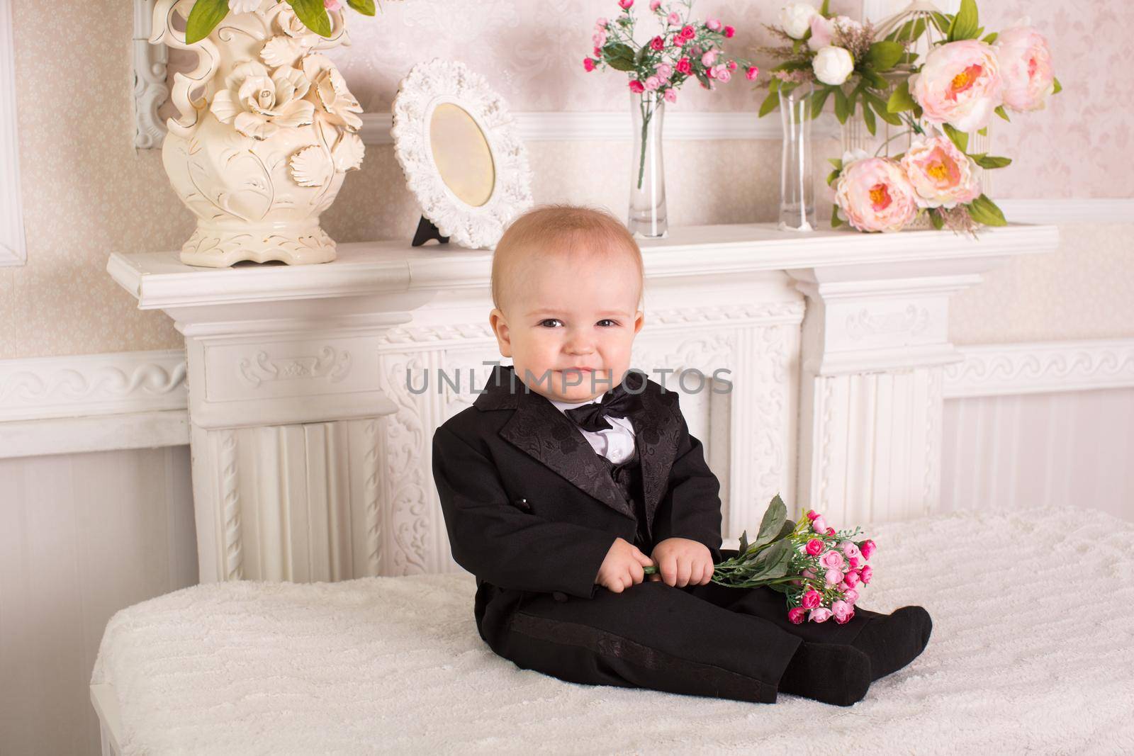Child in a tuxedo sitting on the bed next to the fireplace with flowers in their hands by StudioPeace