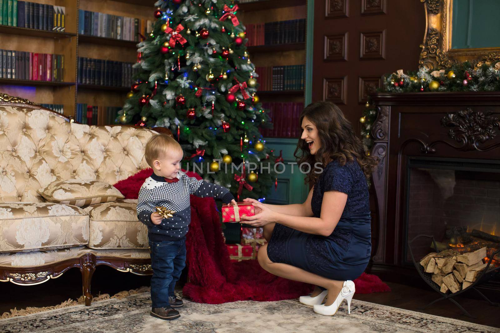 Mother and son open Christmas gifts on the carpet near the Christmas tree and the fireplace.