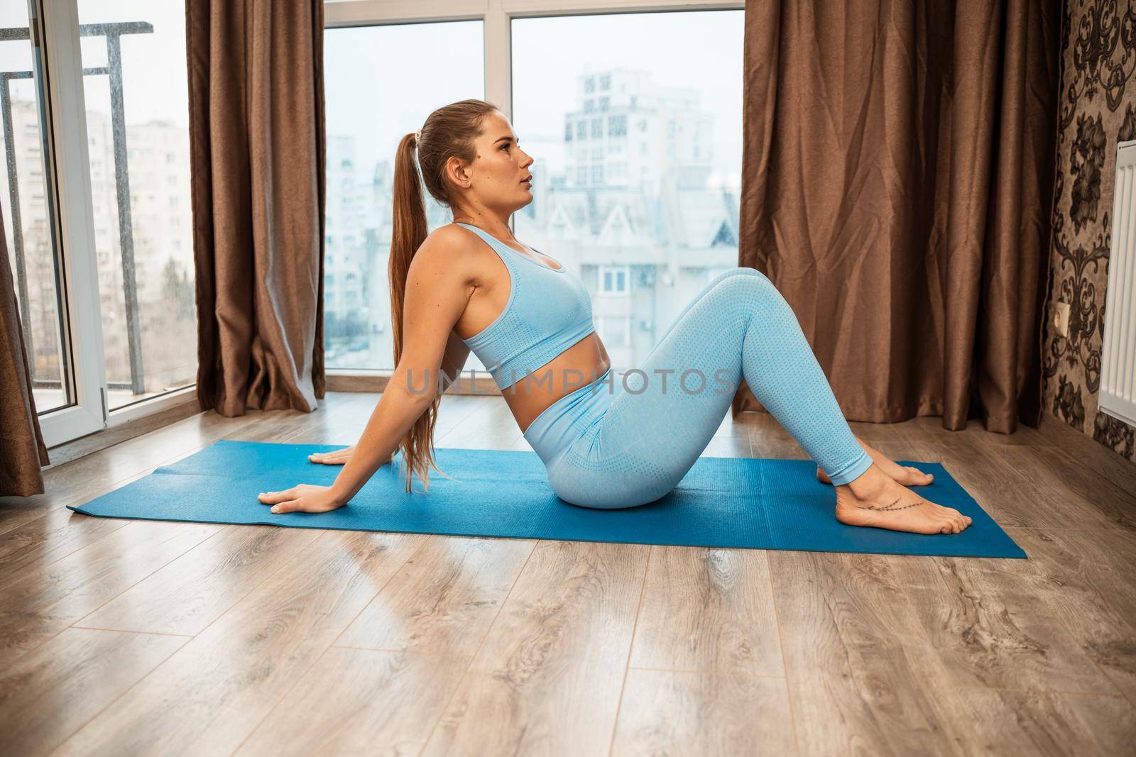 Young athletic attractive woman practicing yoga doing exercises. Works out at home or in a yoga studio, sportswear, blue pants and a full-length top indoors. Healthy lifestyle concept.