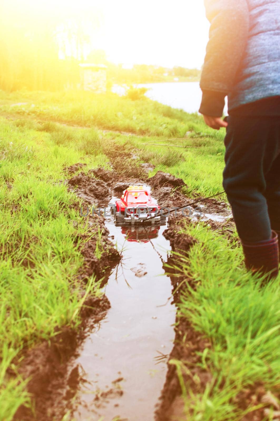 a child in a blue yolk and rubber boots carries a typewriter by a rope through a puddle. A child walks outside after a summer rain. A concept of a carefree childhood.