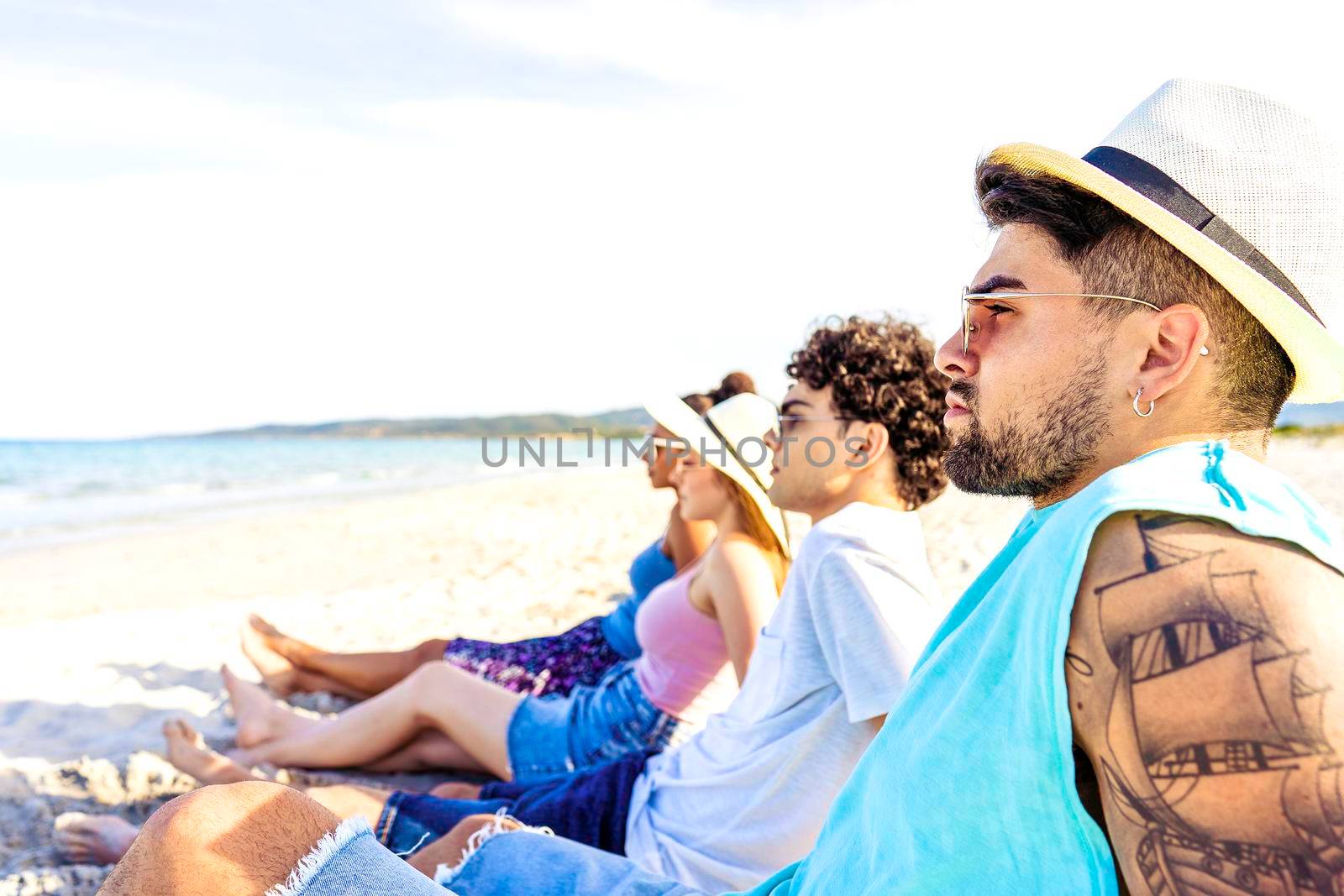Selective focus portrait of handsome hipster guy with tattooed arm in white Panama hat sitting on seashore with friends looking at ocean water in vacation resort. Modern young people enjoying travel by robbyfontanesi