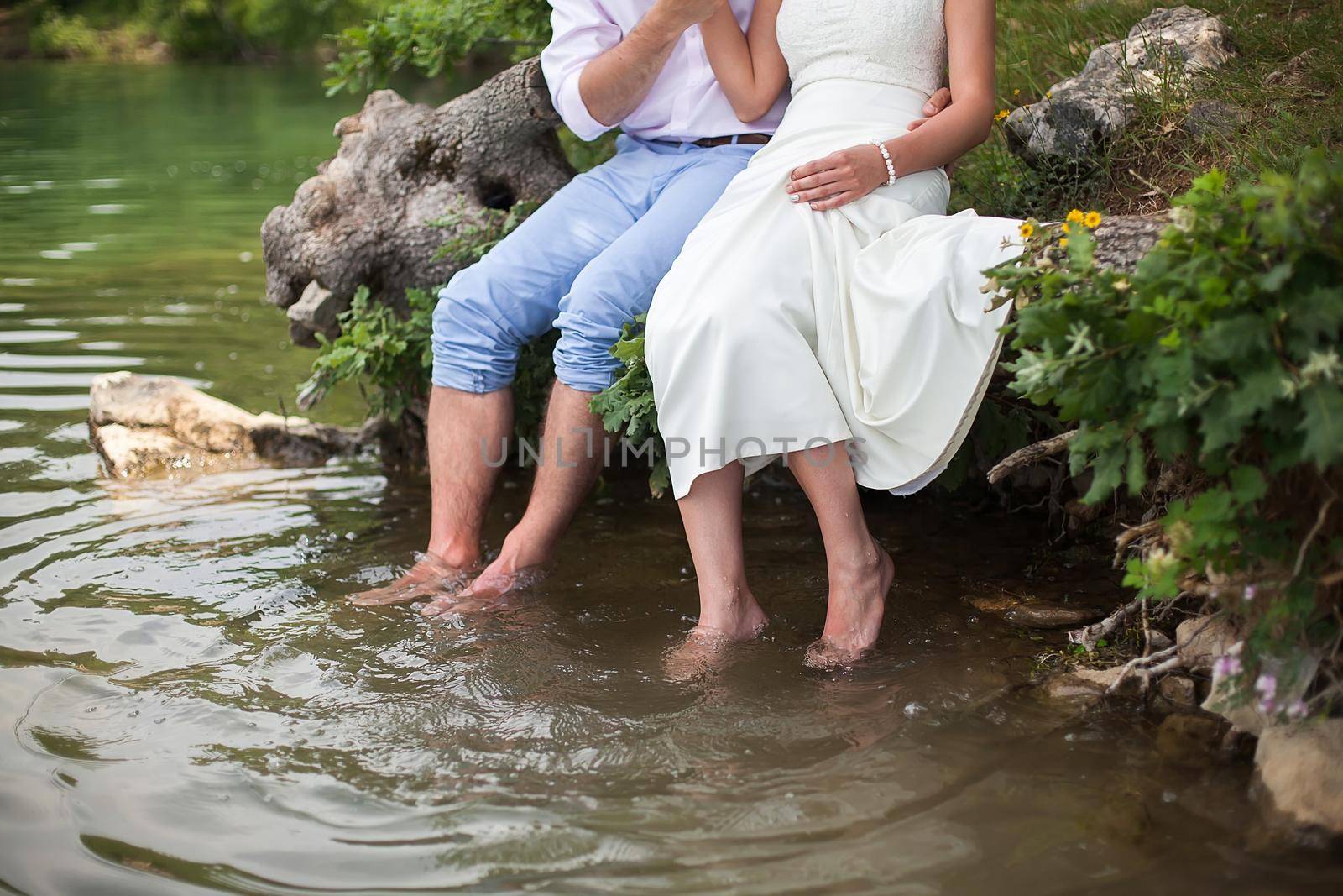 The bride and groom sitting on the shore of the lake. Foot splashing in the water.