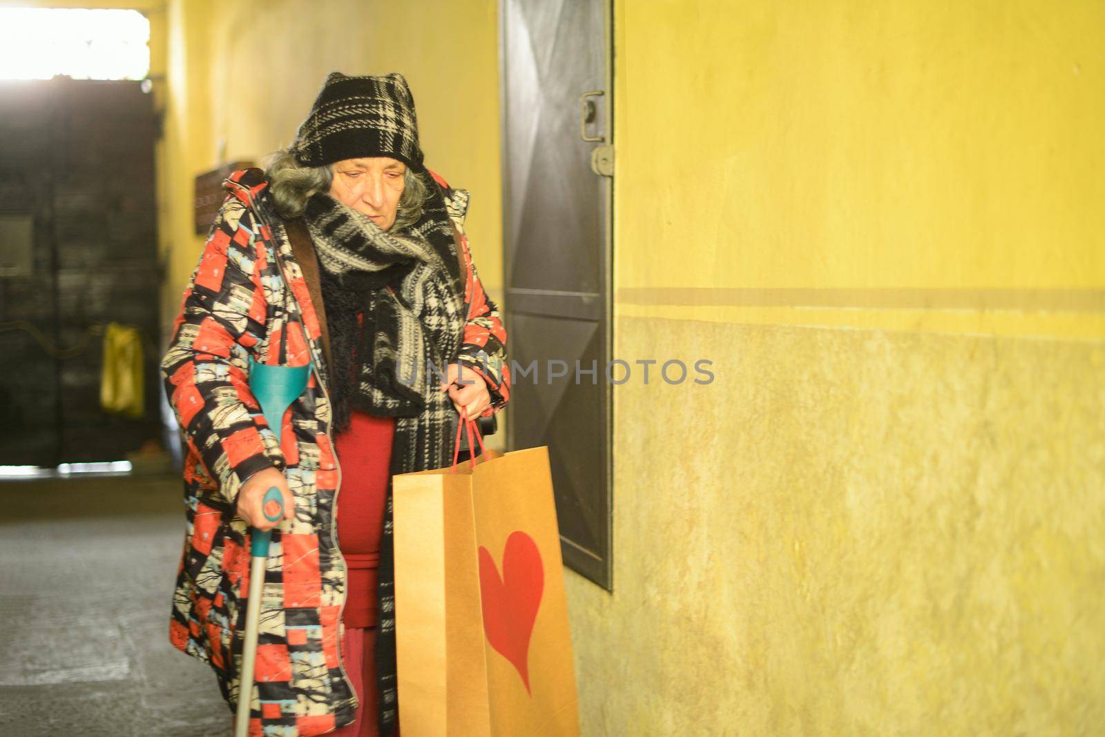 senior east european disabled woman entering a building with an architectural barrier opening a gate with one hand and holdding the crutch with the other also grabbing a bag dressed warm and shabby. High quality photo