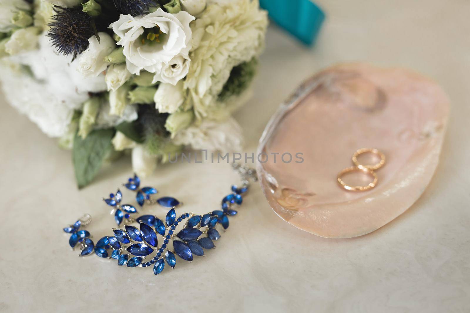 Gold wedding rings on a pink marble shell next to blue jewelry and flowers. by StudioPeace