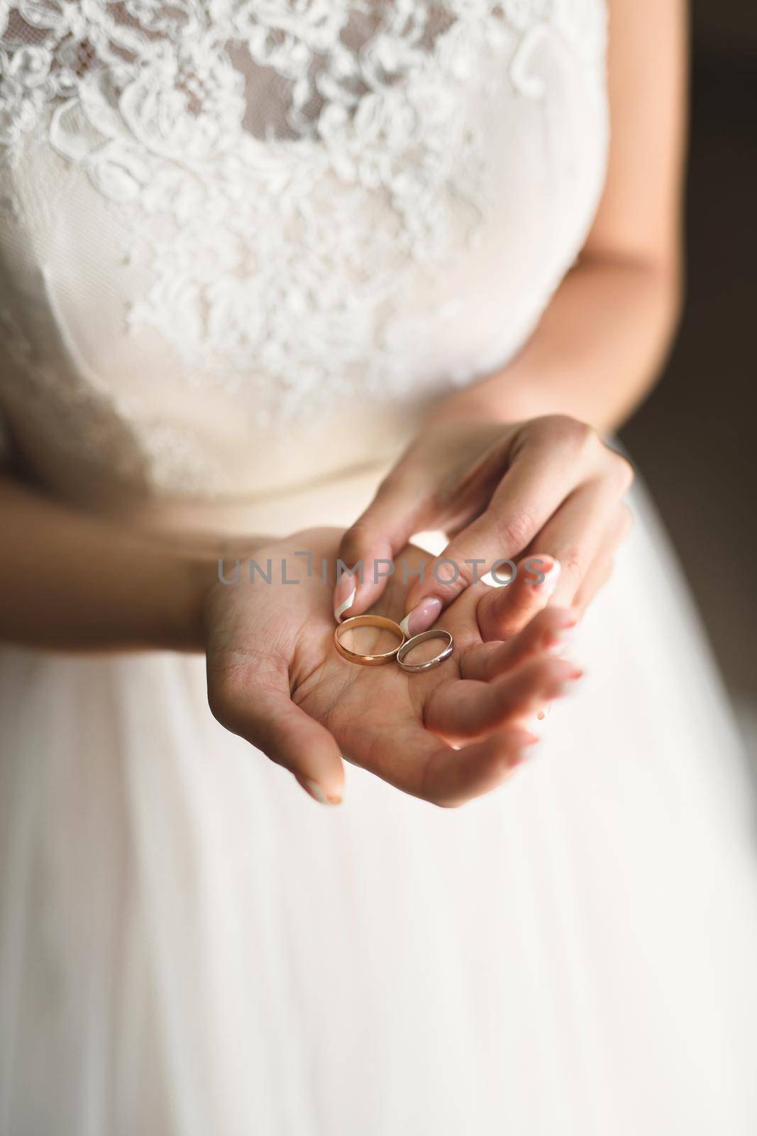 Bride in a white dress holds gold wedding rings in her hands. by StudioPeace