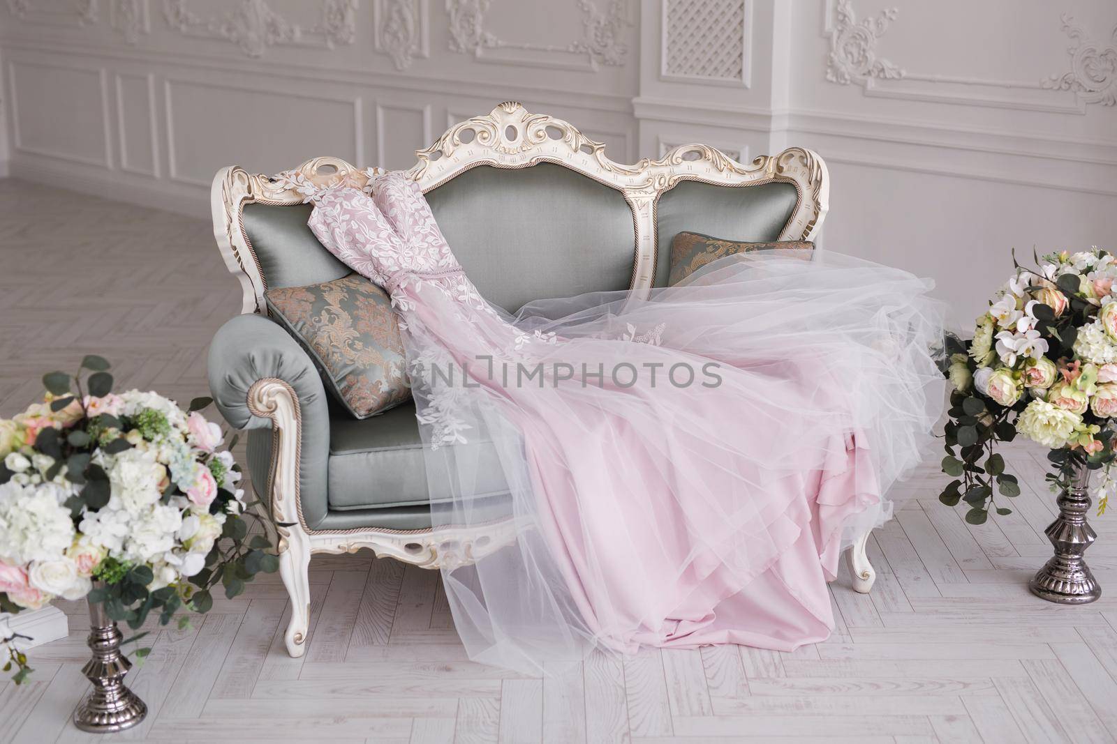 Luxurious pink wedding dress is lying on a grey sofa in a white room. by StudioPeace