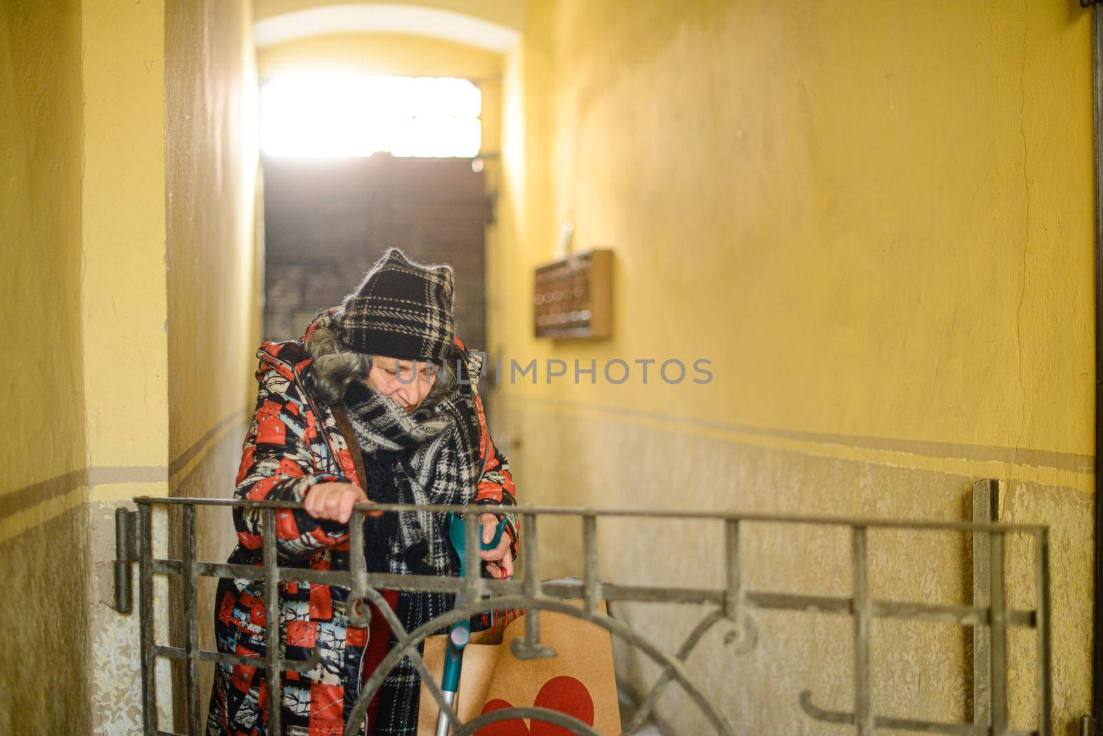 senior east european disabled woman entering a building with an architectural barrier opening a gate with one hand and holdding the crutch with the other also grabbing a bag dressed warm and shabby. High quality photo