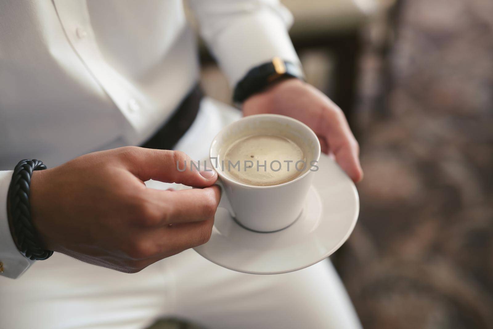 An elegant man in a white suit drinks coffee from a white mug. Hands and mug close -up