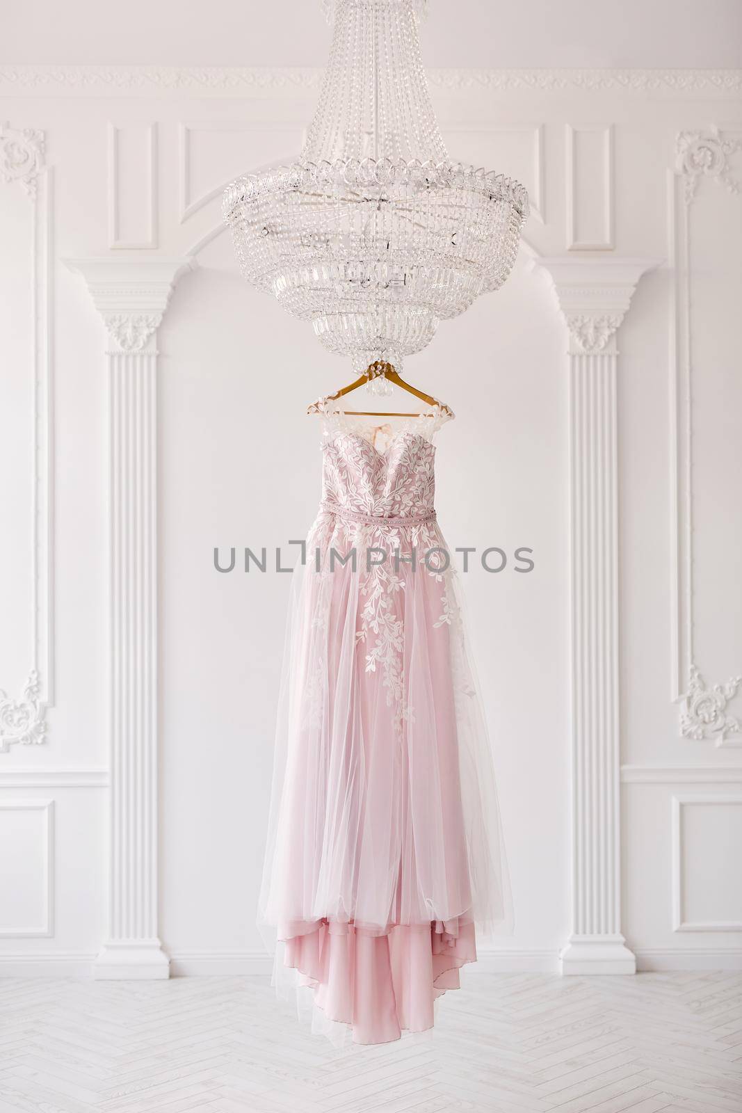 Rich pink wedding dress hangs on a chandelier in a white room. by StudioPeace