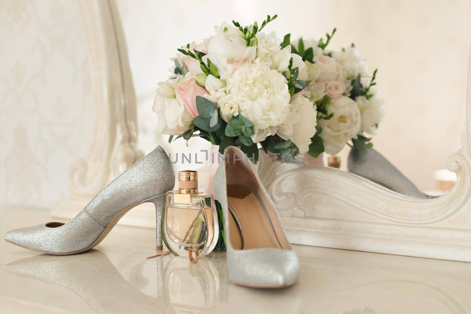 Silver shoes of the bride , perfume, bouquet and wedding rings on the dressing table near the mirror. by StudioPeace