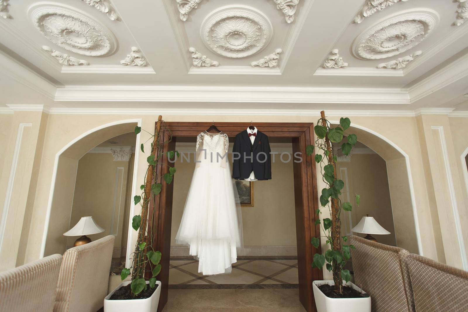 The bride's wedding dress and the groom's jacket on wooden hangers, hang on the wardrobe by StudioPeace