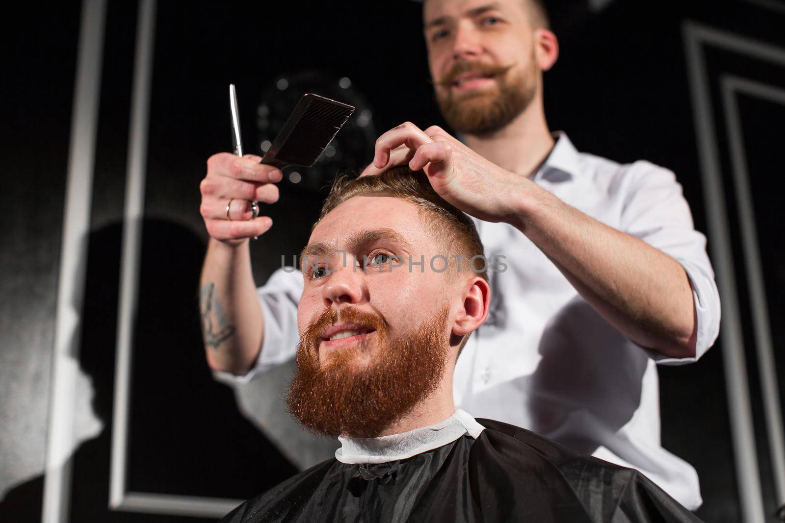 Master cuts hair and beard in the Barber shop. Hairdresser makes hairstyle using scissors and a metal comb. by StudioPeace