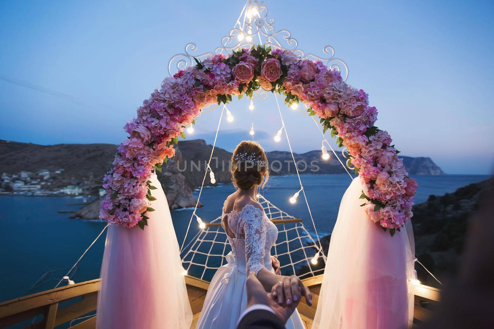 The bride holds the groom's hand against the background of a wedding arch with bright lights at sunset on the seashore. The wedding ceremony.