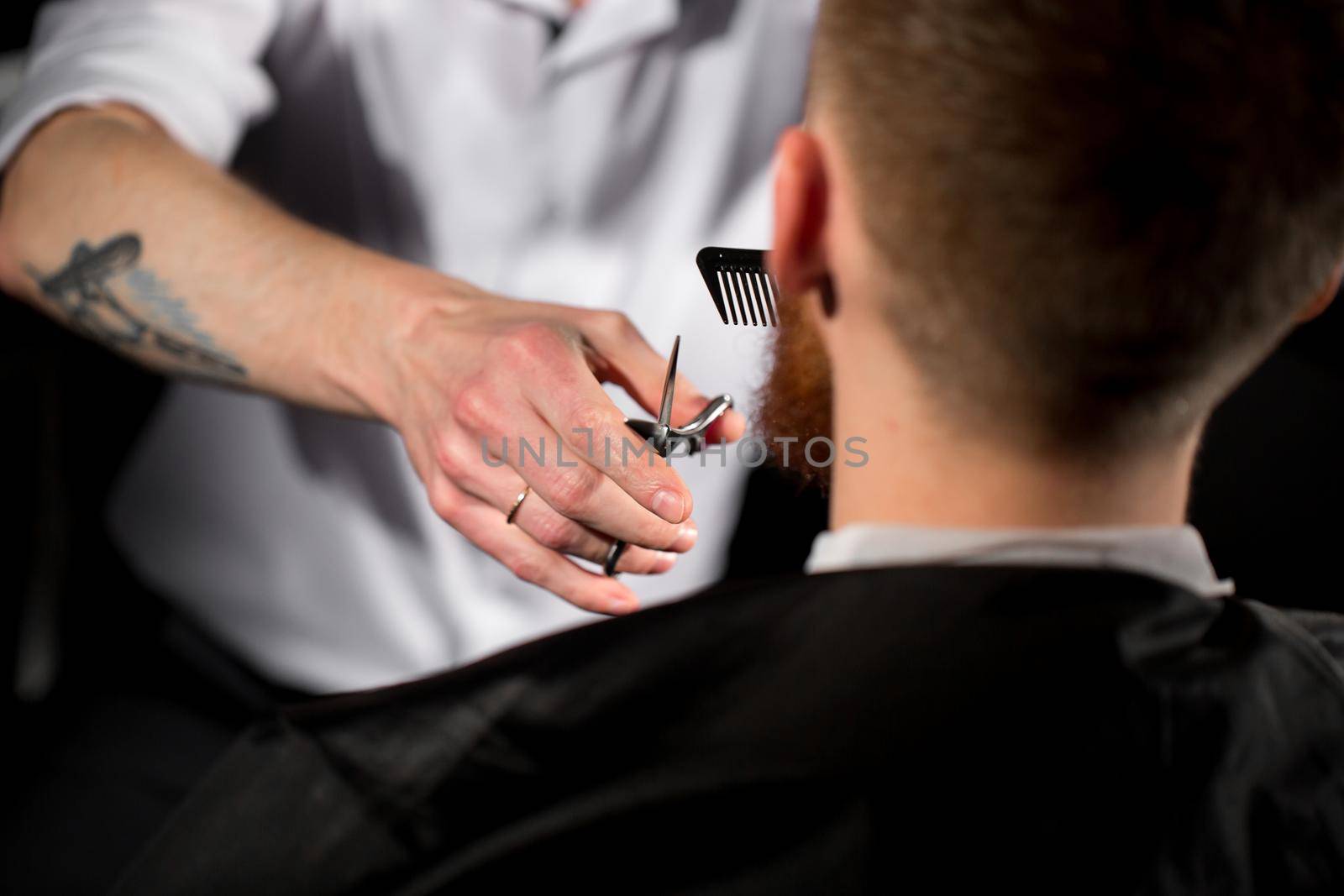 Master cuts hair and beard in the Barber shop. Hairdresser makes hairstyle using scissors and a metal comb.