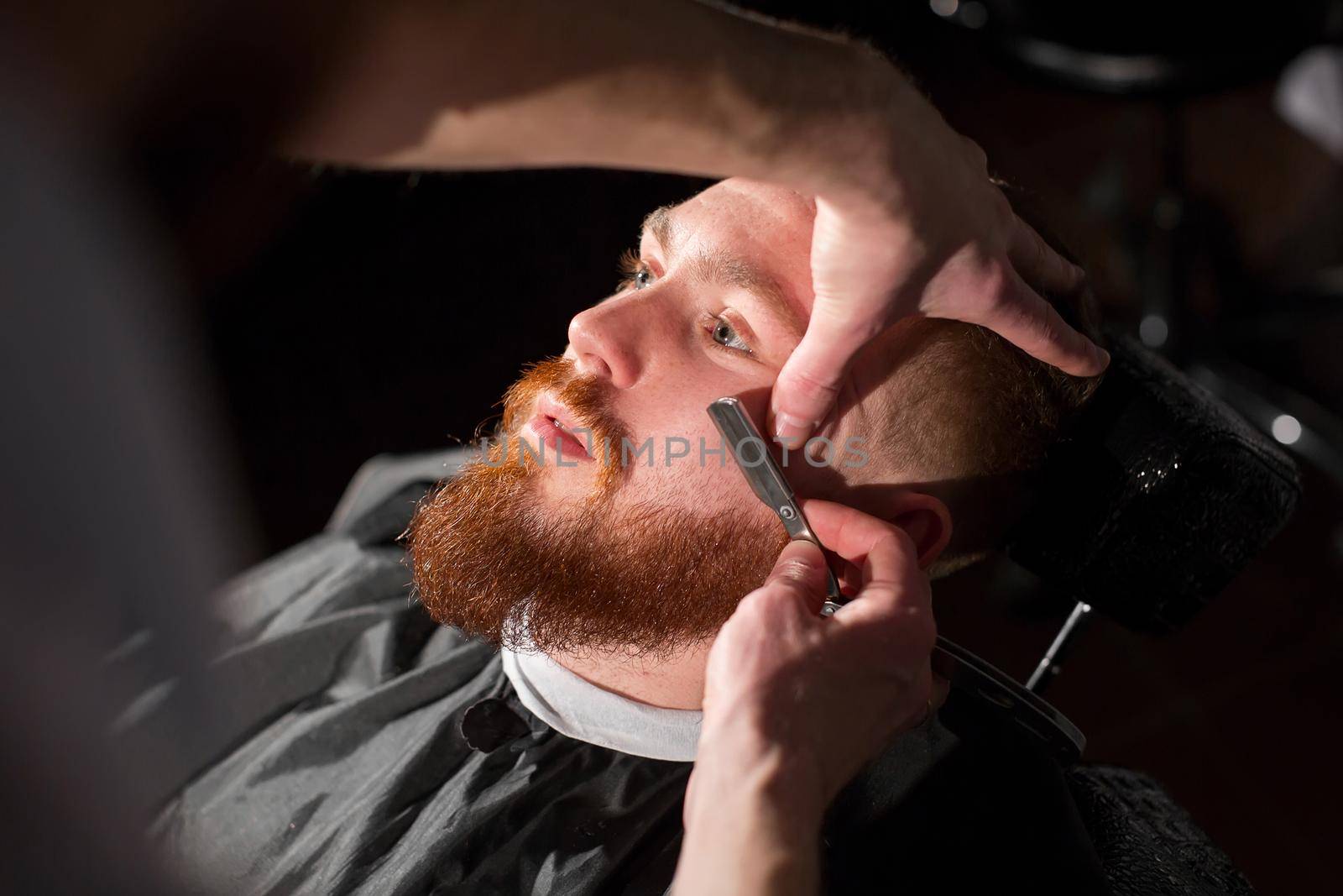 Hairdresser is shaving male beard with the knife. Handsome bearded man is getting shaved by hairdresser at the barbershop. by StudioPeace
