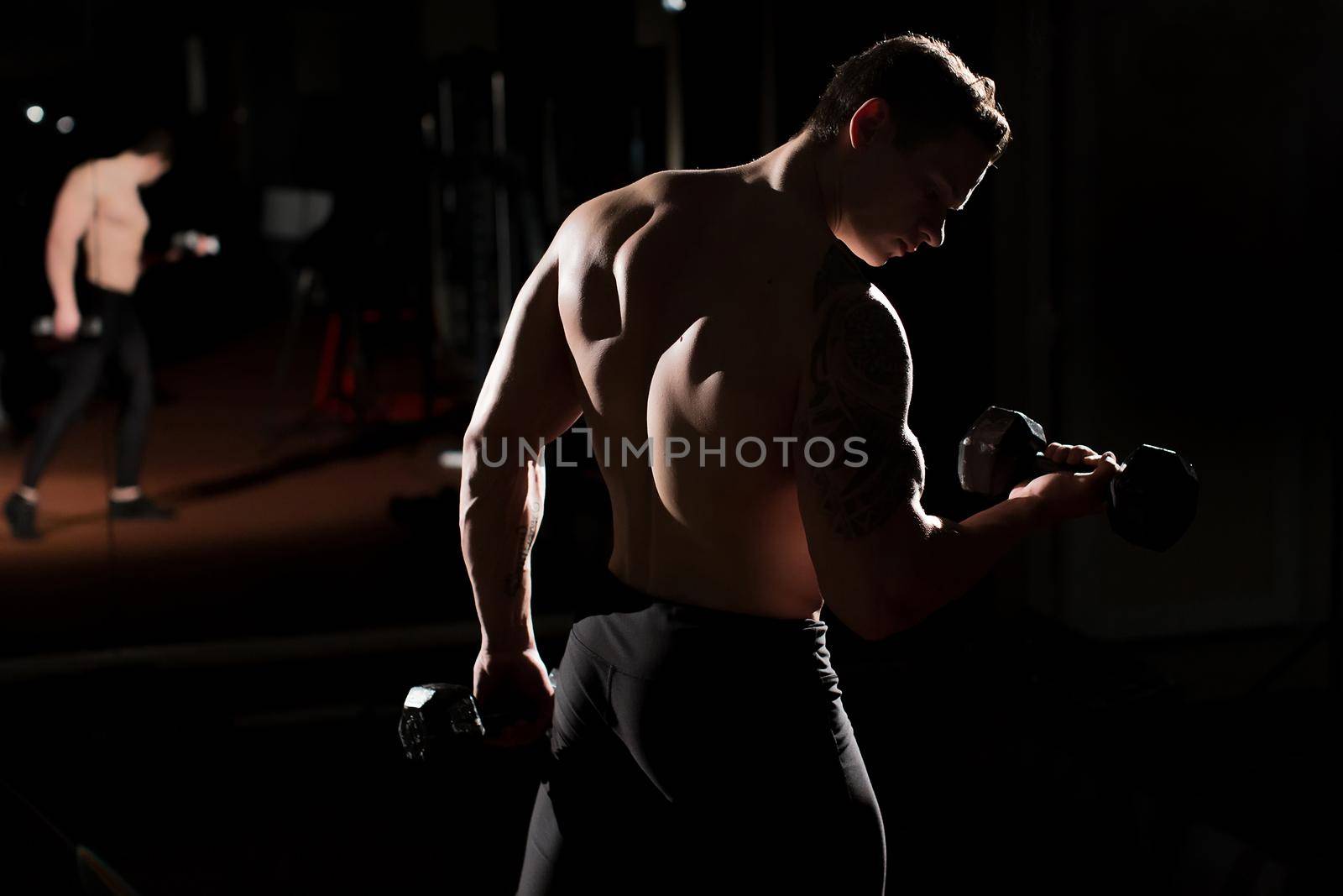 Handsome power athletic guy bodybuilder doing exercises with dumbbell. Fitness muscular body on dark background by StudioPeace