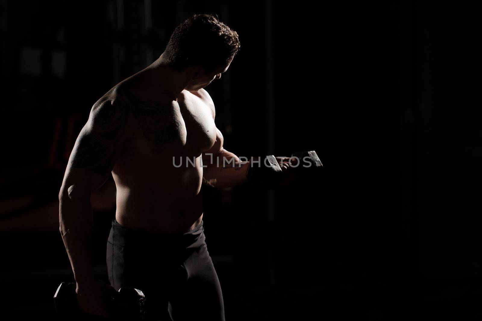 Handsome power athletic guy bodybuilder doing exercises with dumbbell. Fitness muscular body on dark background.