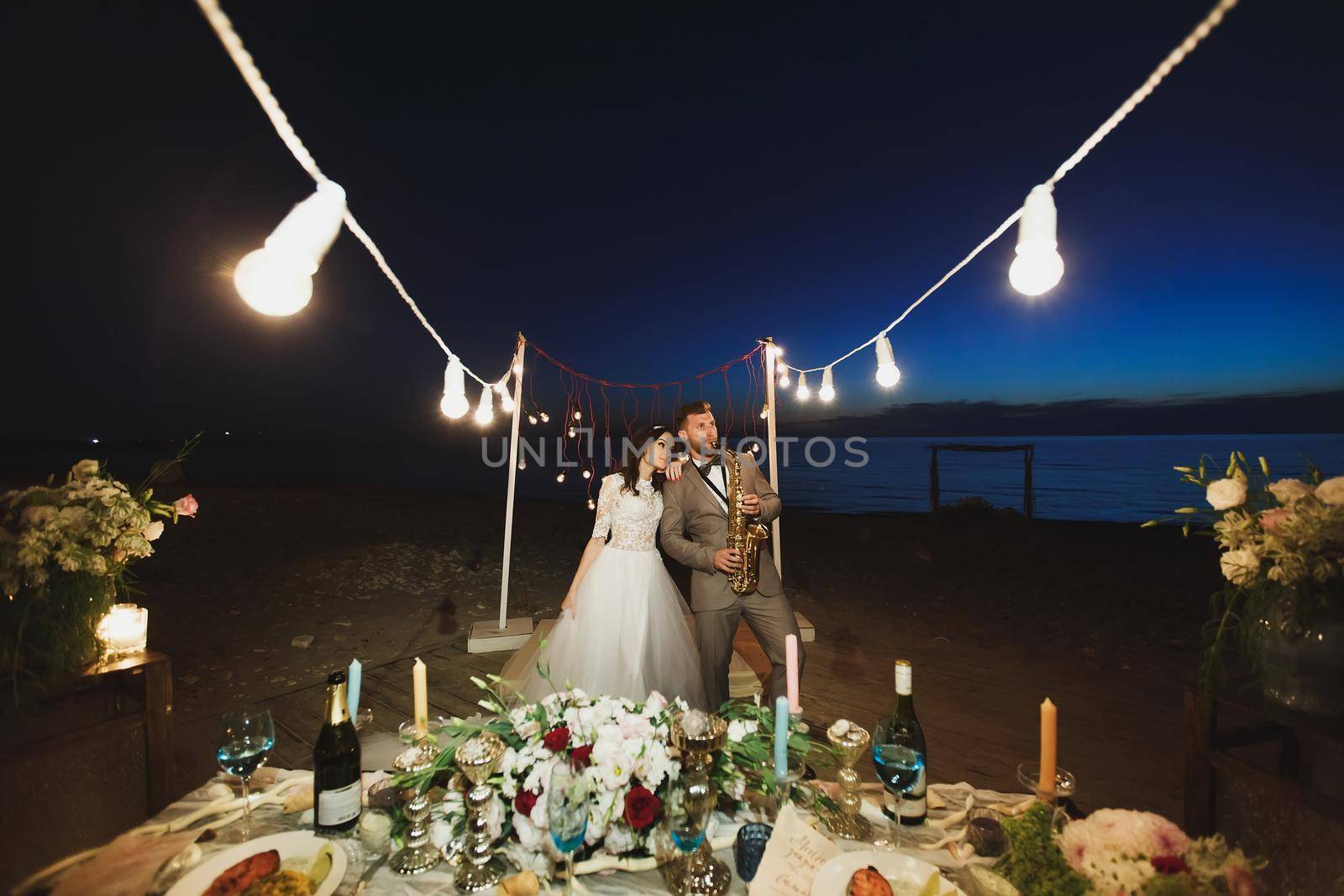 Wedding banquet on the ocean shore at night. The groom is playing the saxophone by StudioPeace