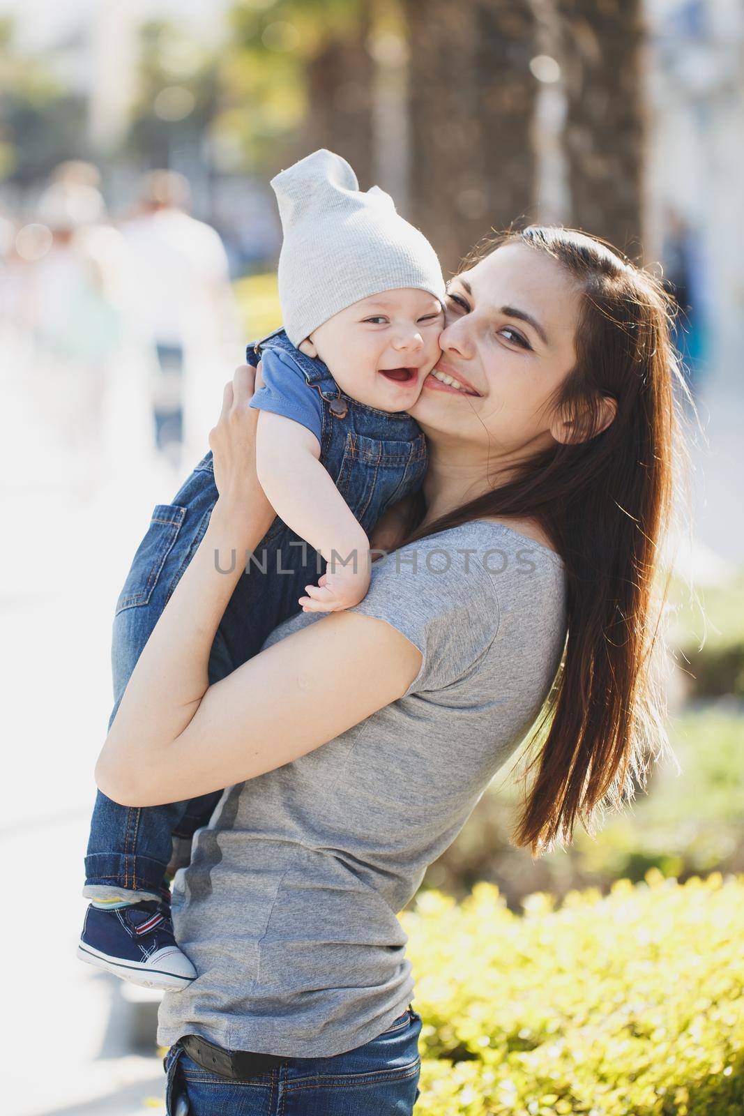 Cute stylish young mother lady kissing cheek nicely dressed son and enjoying nice sunny day. Son on mother hands and smiling by StudioPeace