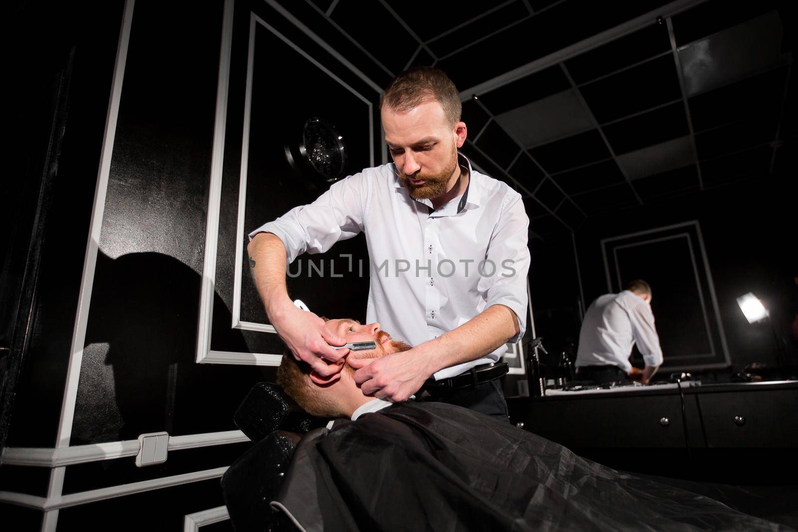 Hairdresser is shaving male beard with the knife. Handsome bearded man is getting shaved by hairdresser at the barbershop