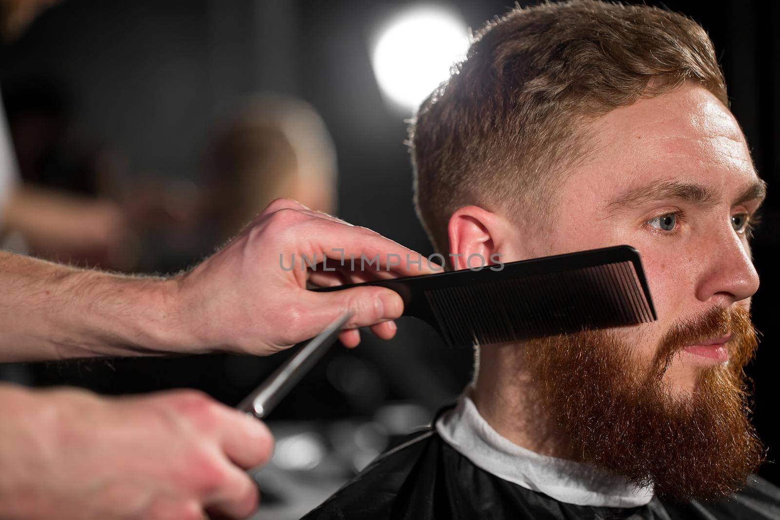 Master cuts hair and beard in the Barber shop. Hairdresser makes hairstyle using scissors and a metal comb. by StudioPeace