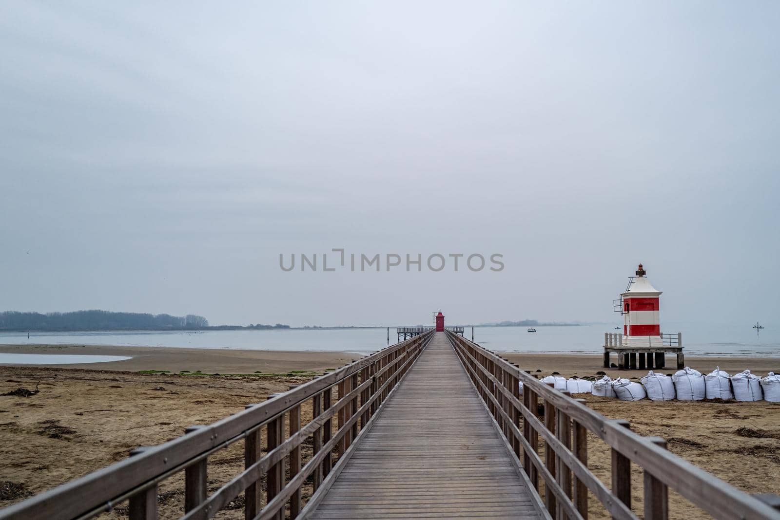 Small red and white wooden lighthouse on a wintry sandy beach next to a wooden jetty without people.
