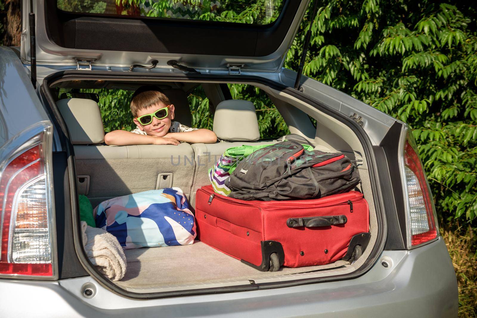 Adorable kid boy wearing sunglasses sitting in car trunk. Portrait of Happy child with open car boot while waiting for parent get ready for vocation. Family trip traveling by car concept.