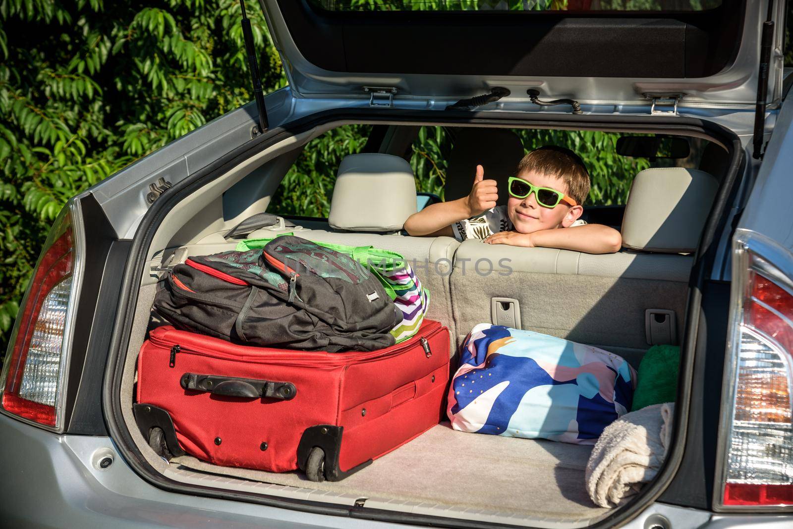 Adorable kid boy wearing sunglasses sitting in car trunk. Portrait of Happy child with open car boot while waiting for parent get ready for vocation. Family trip traveling by car concept.