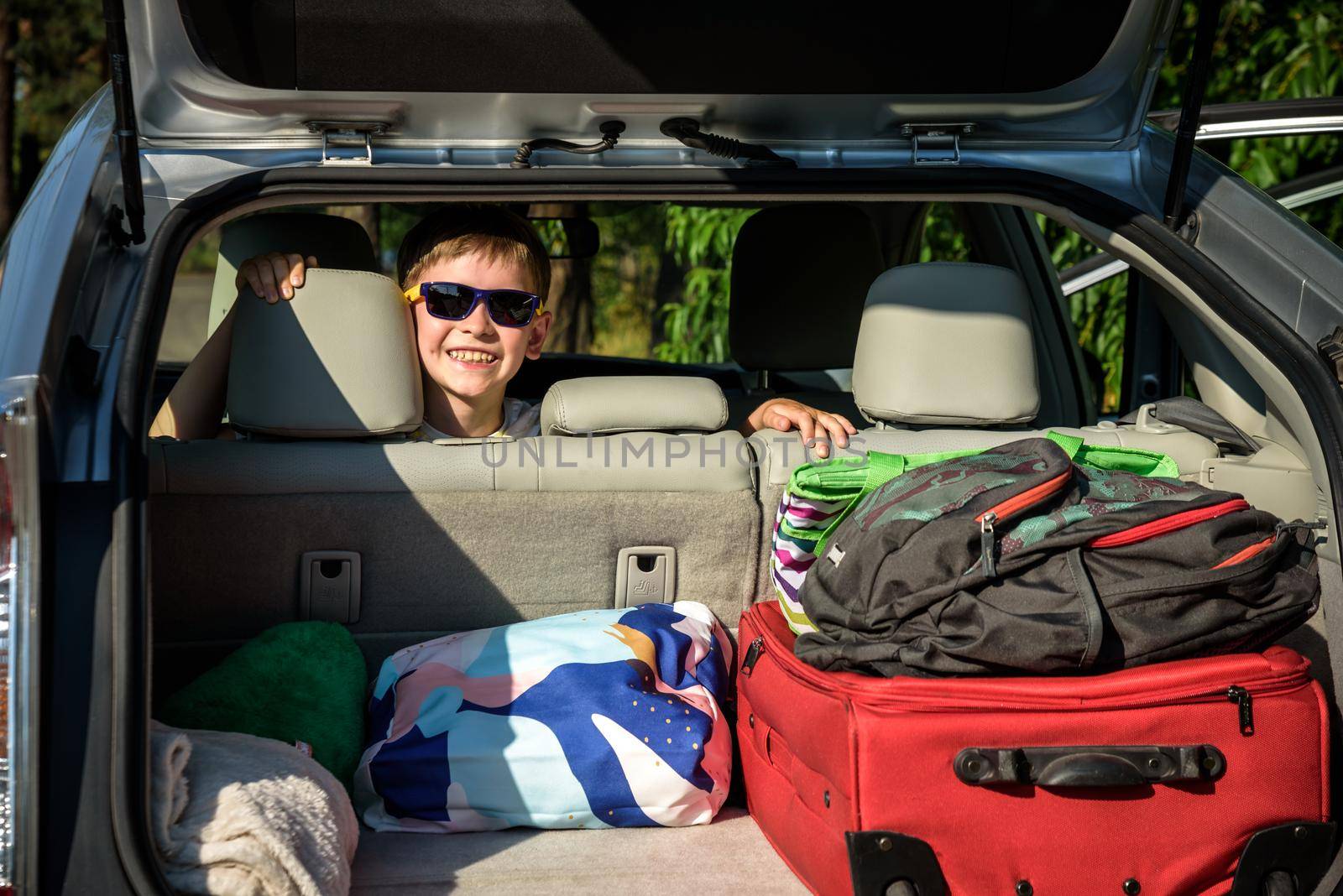 Adorable kid boy wearing sunglasses sitting in car trunk. Portrait of Happy child with open car boot while waiting for parent get ready for vocation. Family trip traveling by car concept by Kobysh