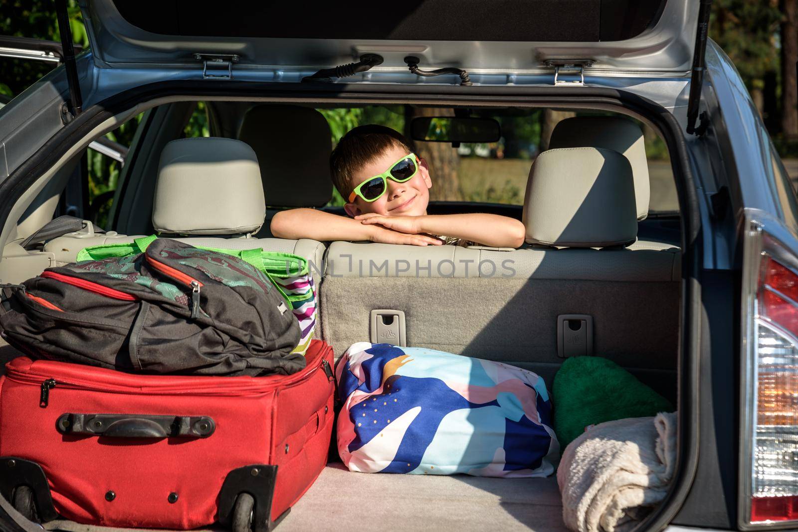 Adorable kid boy wearing sunglasses sitting in car trunk. Portrait of Happy child with open car boot while waiting for parent get ready for vocation. Family trip traveling by car concept by Kobysh