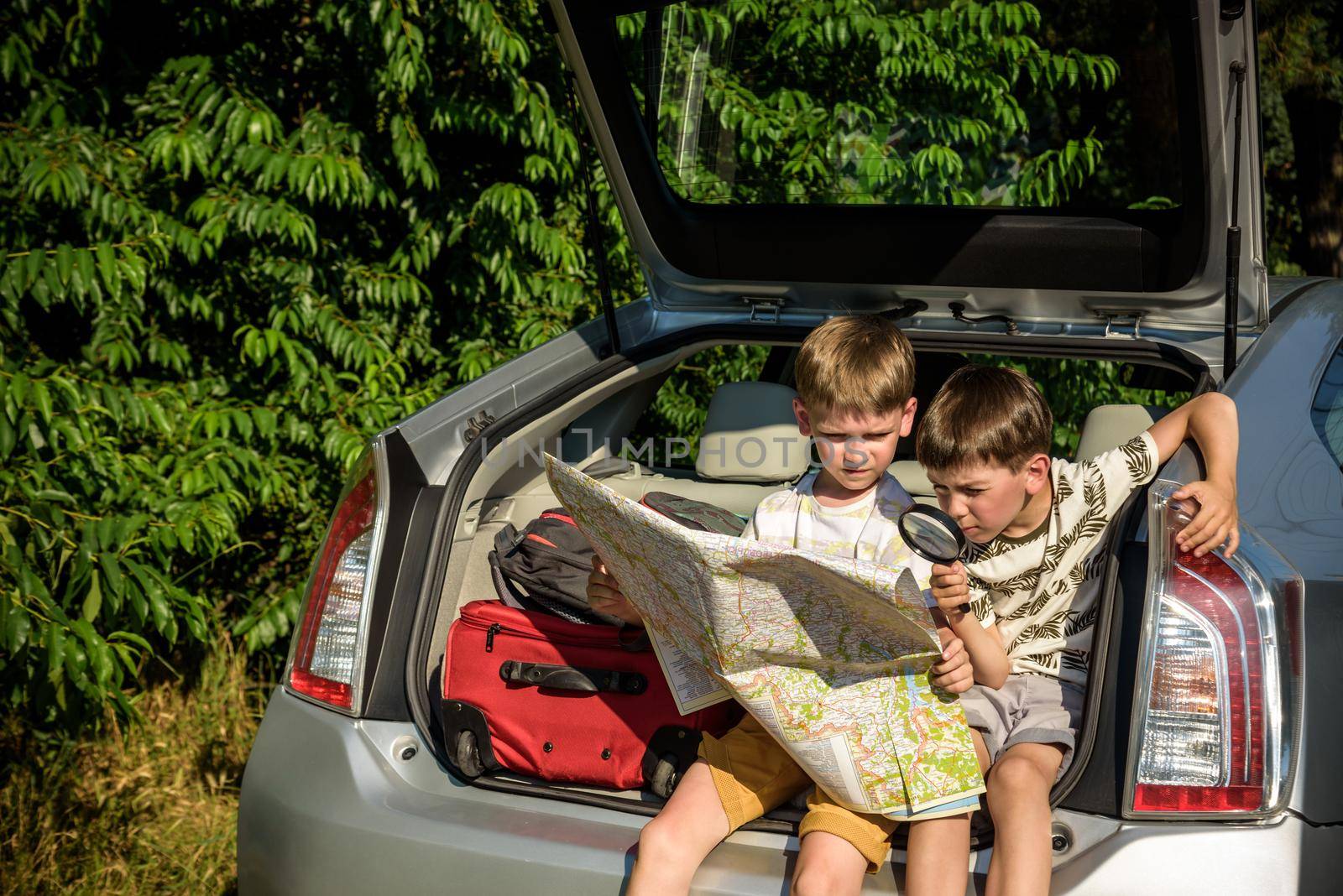 Two cute boys sitting in a car trunk before going on vacations with their parents. Kids sitting in a car examining a map. Summer break at school. Family travel by car.