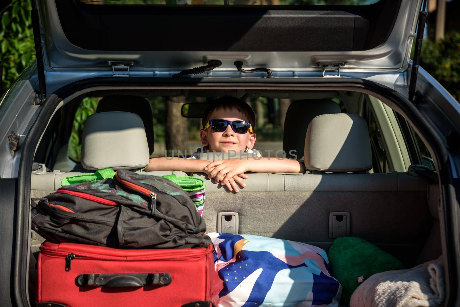 Adorable kid boy wearing sunglasses sitting in car trunk. Portrait of Happy child with open car boot while waiting for parent get ready for vocation. Family trip traveling by car concept.