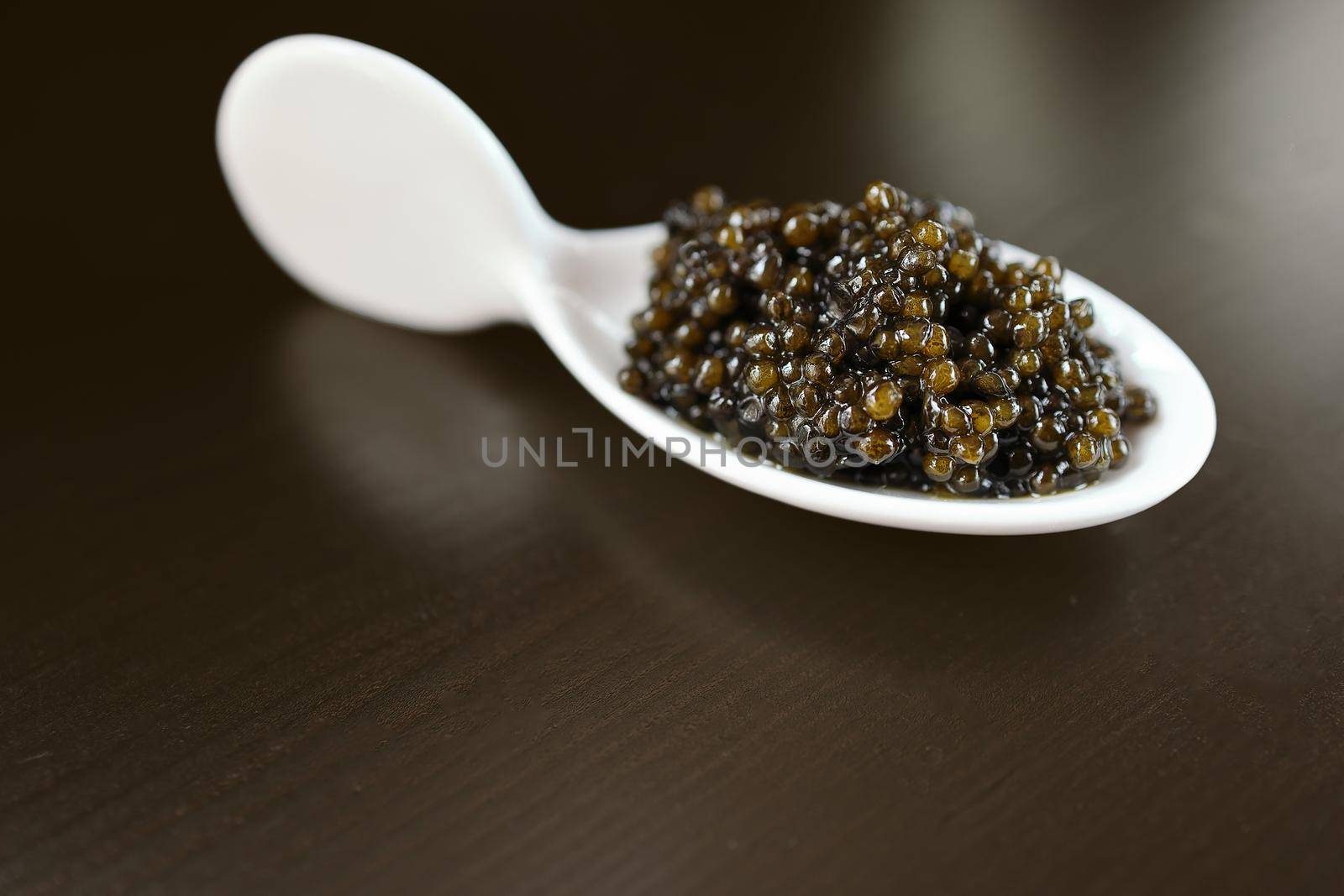 Natural black caviar in a white serving porcelain spoon on a dark background. Selective focus. Minimalistic photo. Daylight. Copy space