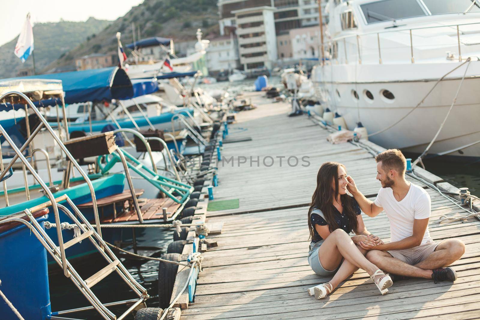 Lovers, guy and girl, are sitting on a wooden pier, holding hands and laughing by StudioPeace