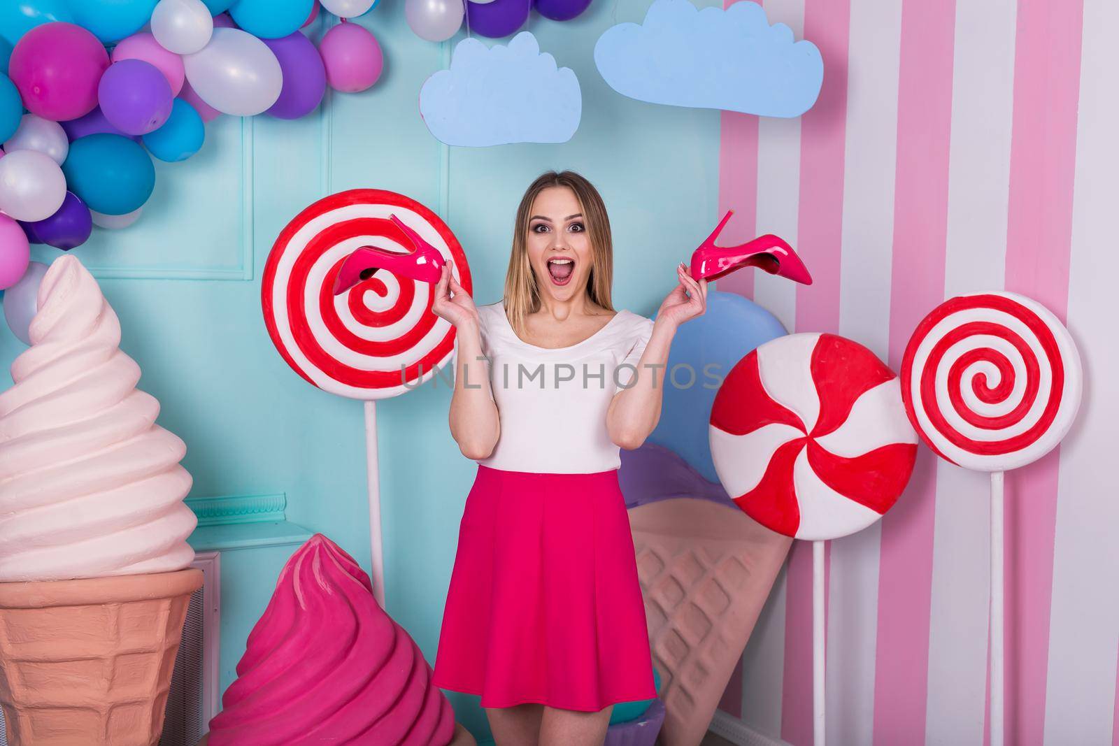 Portrait of joyful young woman in pink dress on background decorated with huge candies and ice cream. Selects red shoes. by StudioPeace