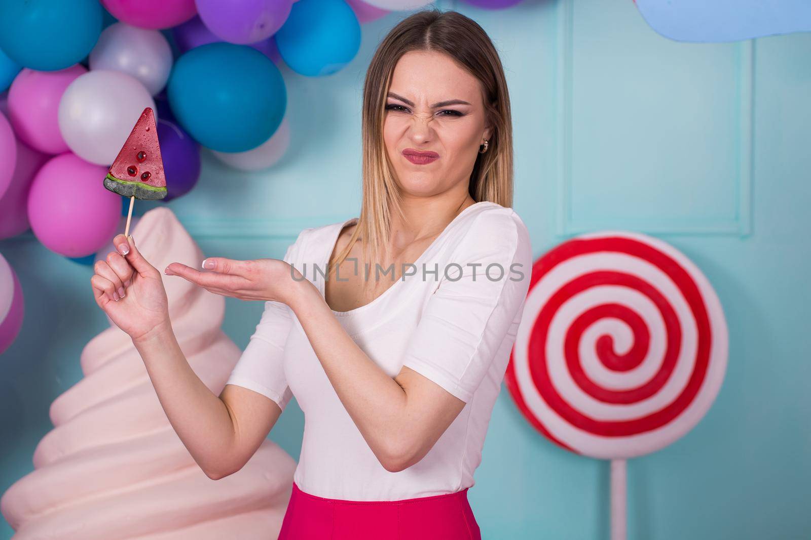 Portrait of amazing sweet-tooth woman in pink dress holding candies and posing on background decorated with huge ice cream. Lollipop watermelon