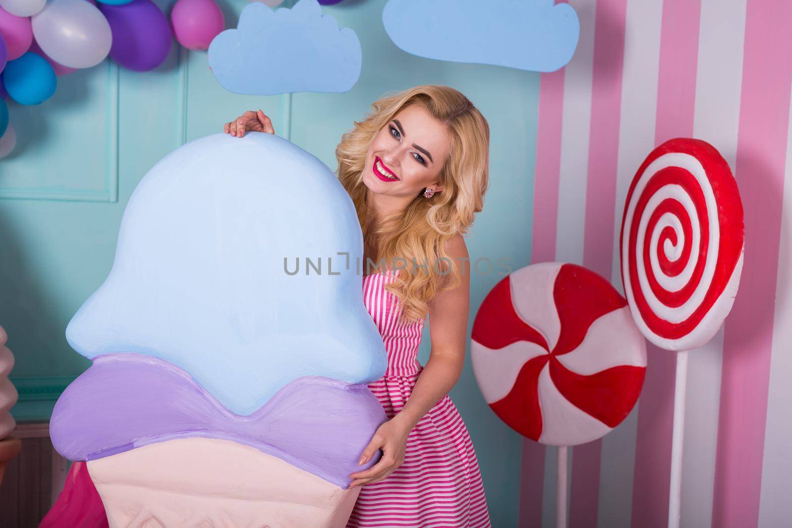 Portrait of young woman in pink dress holding big ice cream and posing on decorated background. Amazing sweet-tooth girl surrounded by toy sweets by StudioPeace