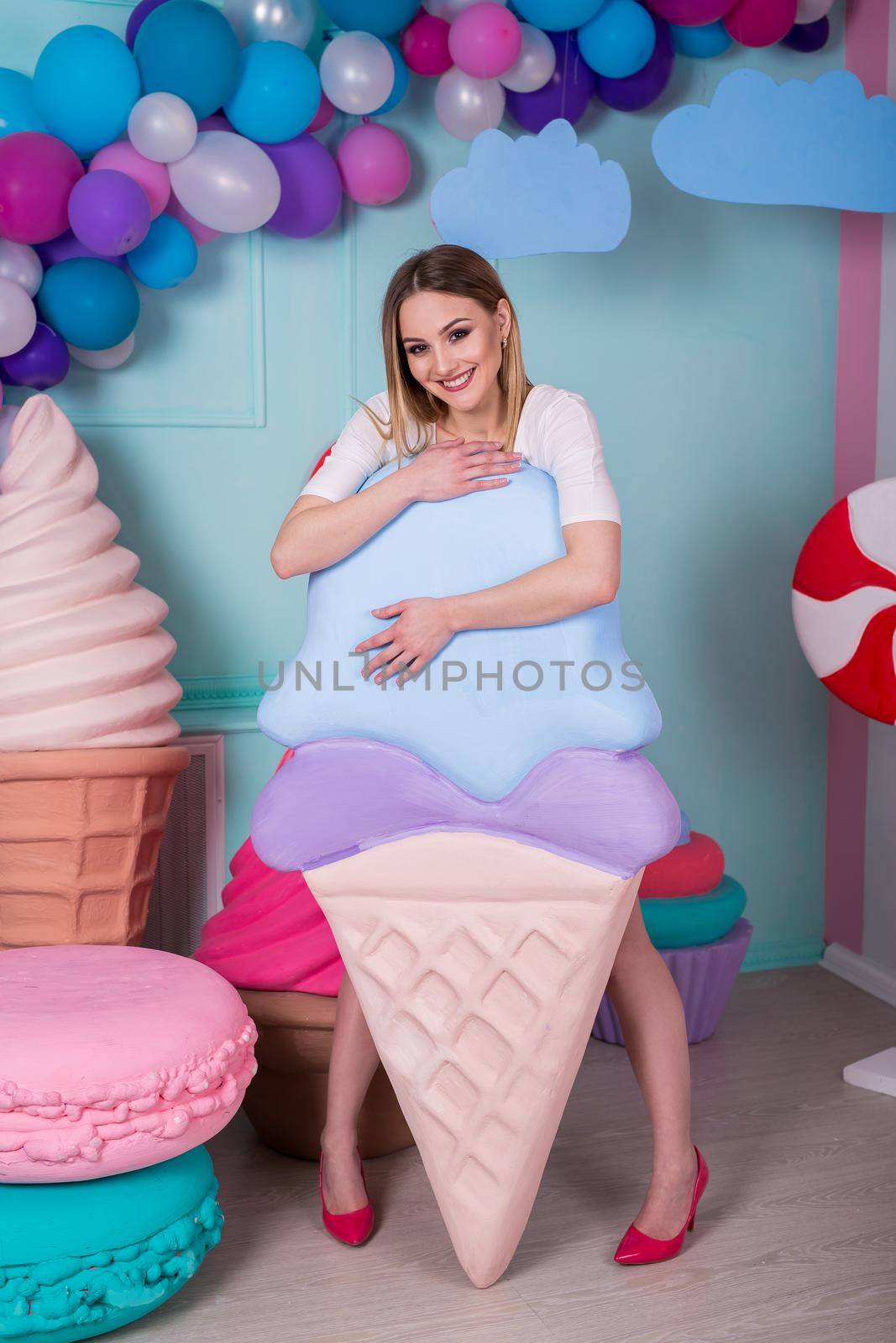 Portrait of young woman in pink dress holding big ice cream and posing on decorated background. Amazing sweet-tooth girl surrounded by toy sweets by StudioPeace