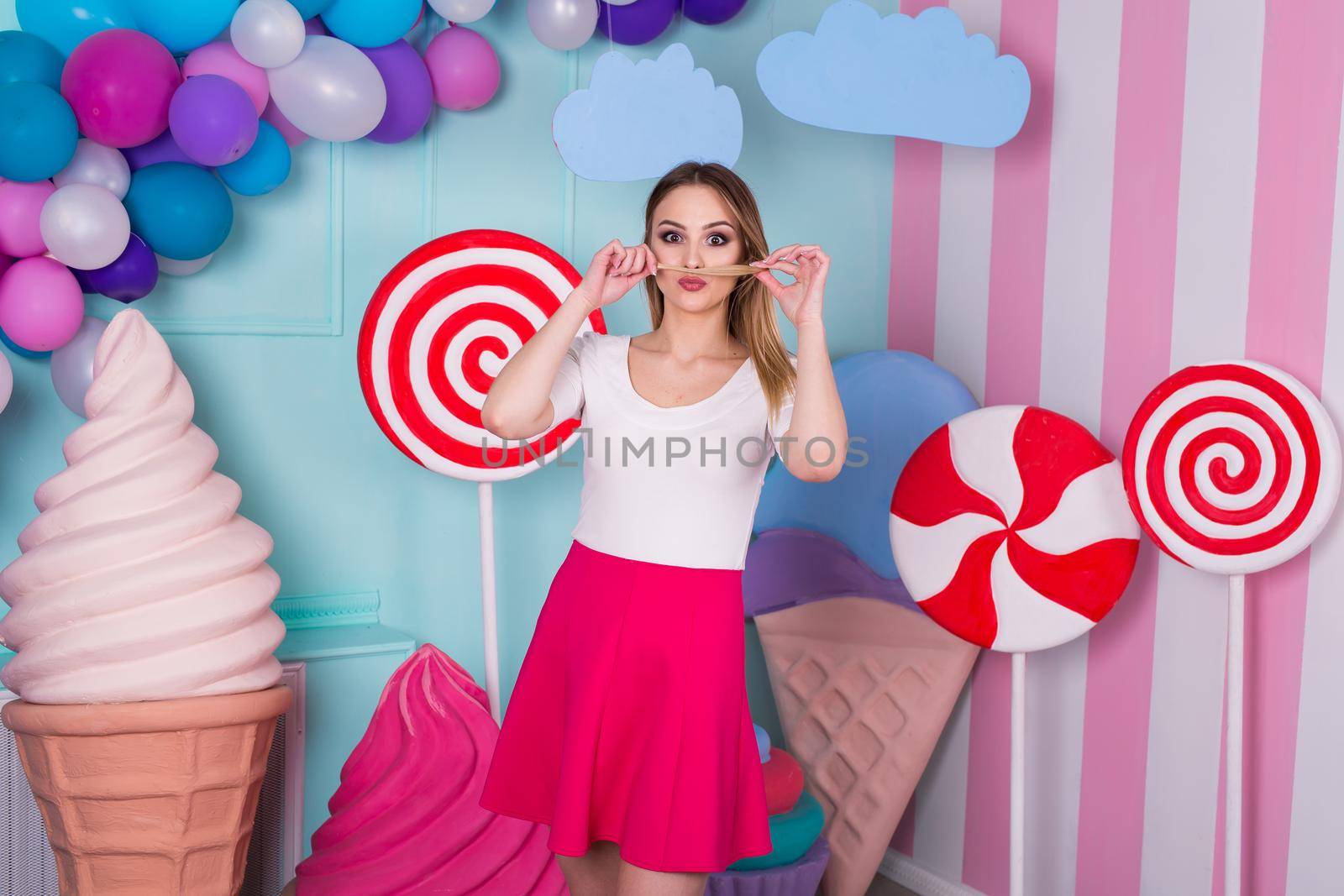 Portrait of joyful young woman in pink dress on background decorated with huge candies and ice cream.
