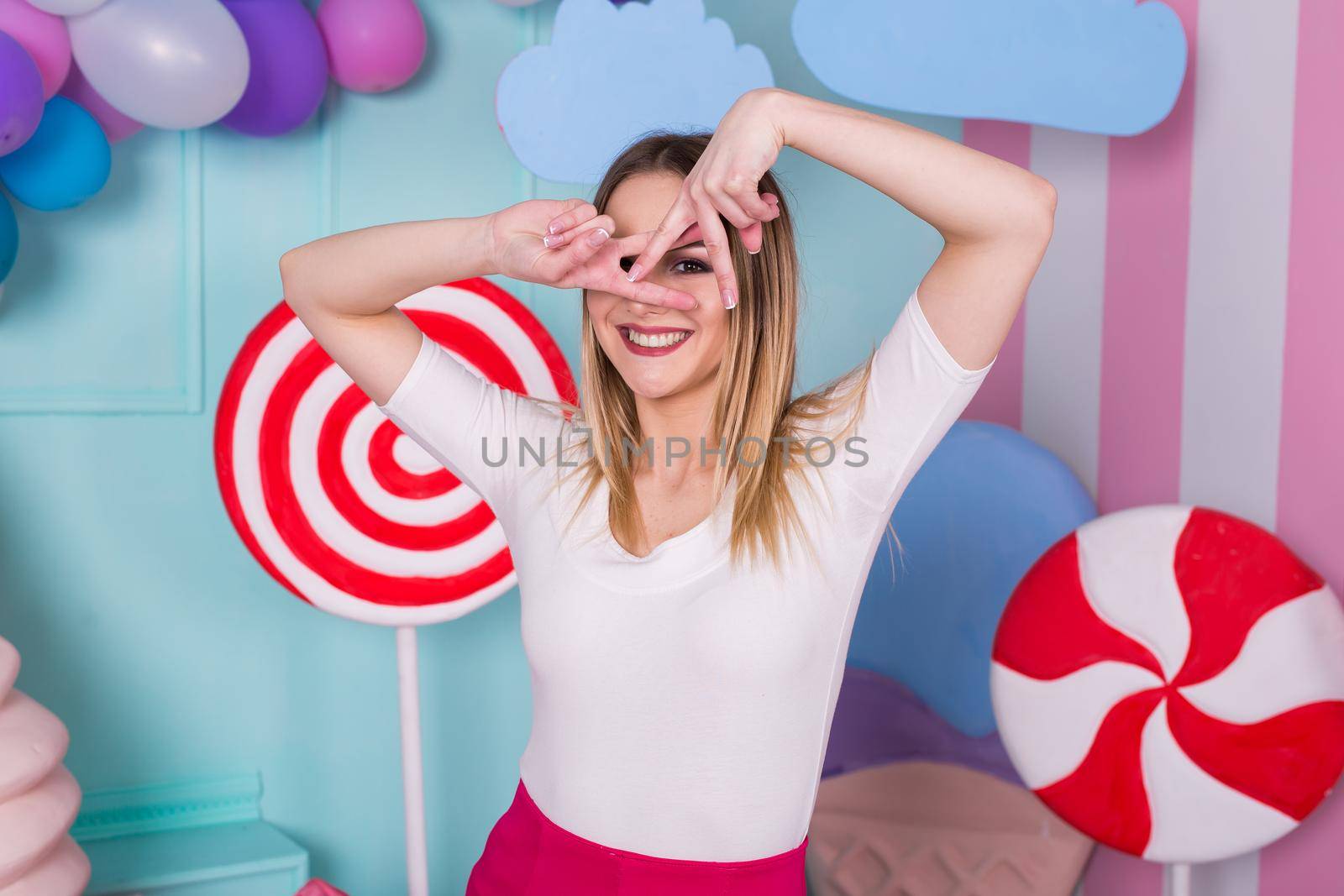 Portrait of joyful young woman in pink dress on background decorated with huge candies and ice cream.