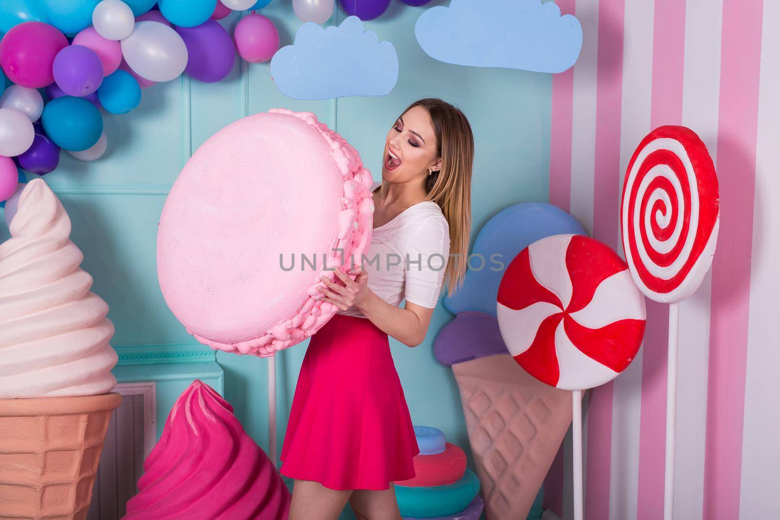 Portrait of young woman in pink dress holding big macaroon and posing on decorated background. Amazing sweet-tooth girl surrounded by toy sweets by StudioPeace