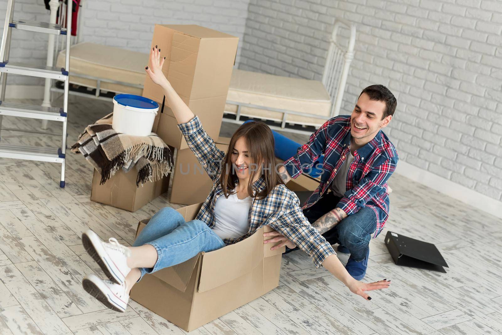 Happy couple having fun laughing moving into new home, young excited woman riding sitting in cardboard box while man pushing it, cheerful roommates playing while packing unpacking belongings together. by StudioPeace