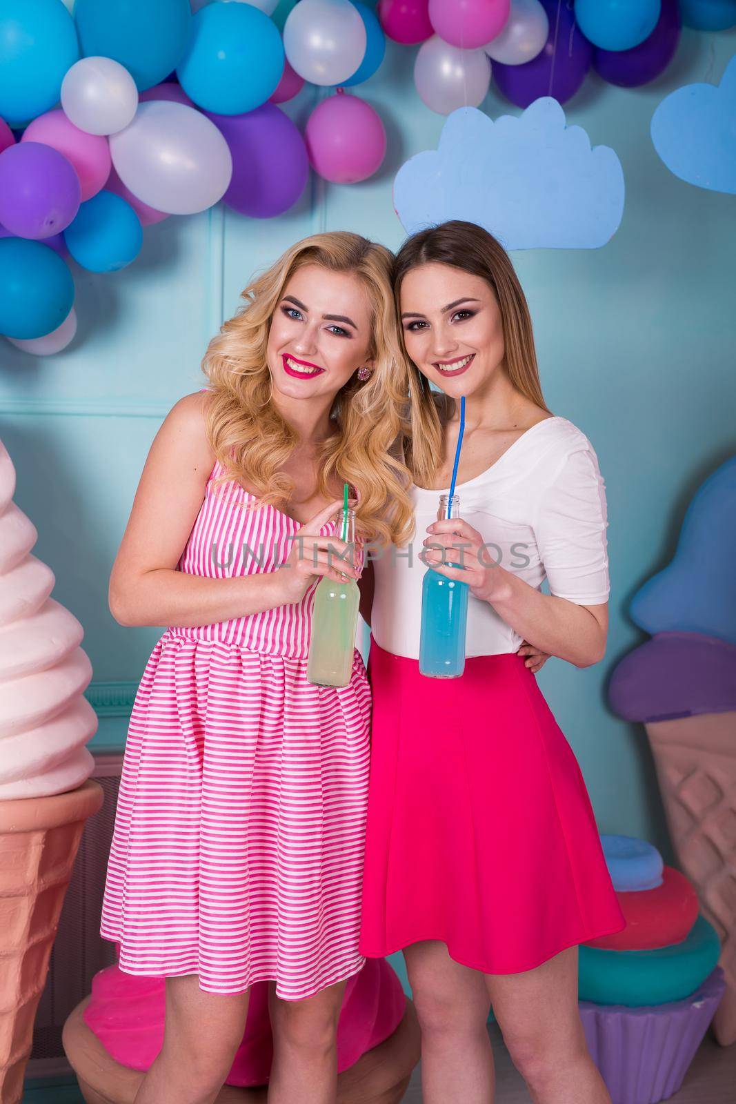 Two young women in pink dresses drink juice in a glass bottle.