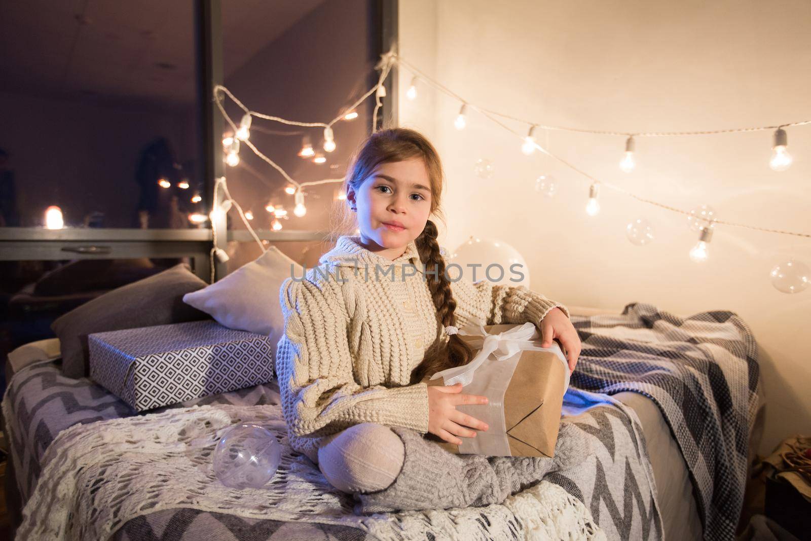 Beautiful little girl sitting on bed with a gift in its hands. Deploy Christmas gift box on holiday in the evening in the beautiful interior room. New year.