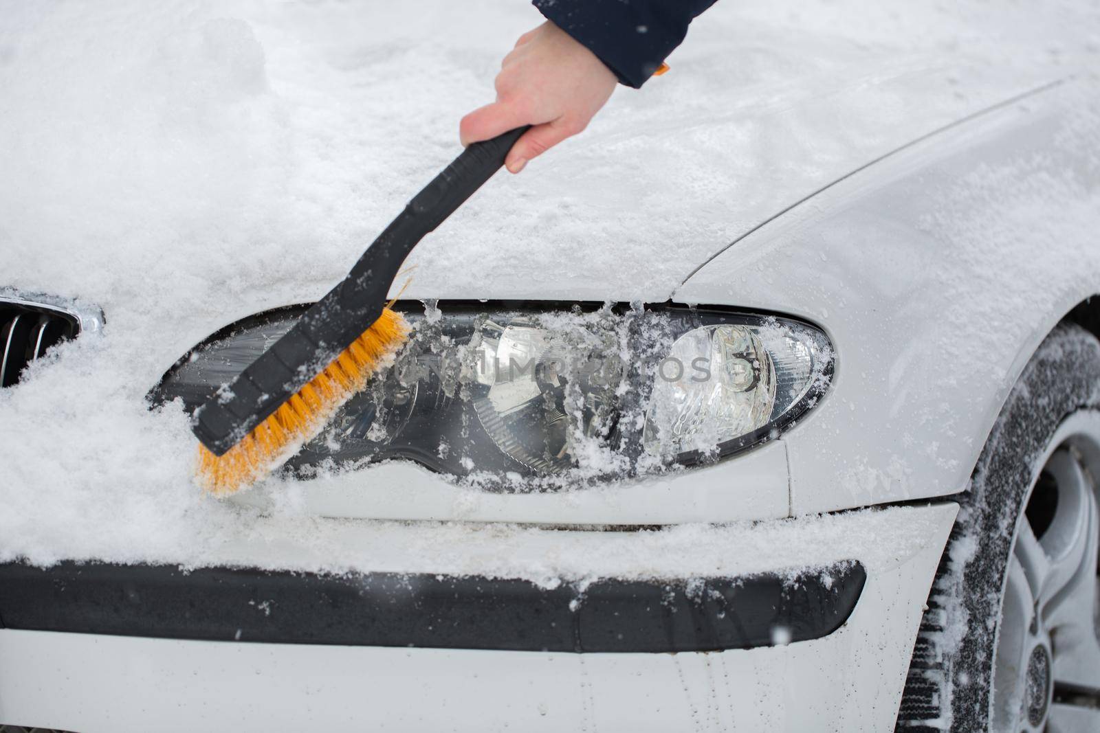 Woman cleaning snow from the car in the winter. Headlight of the car by StudioPeace