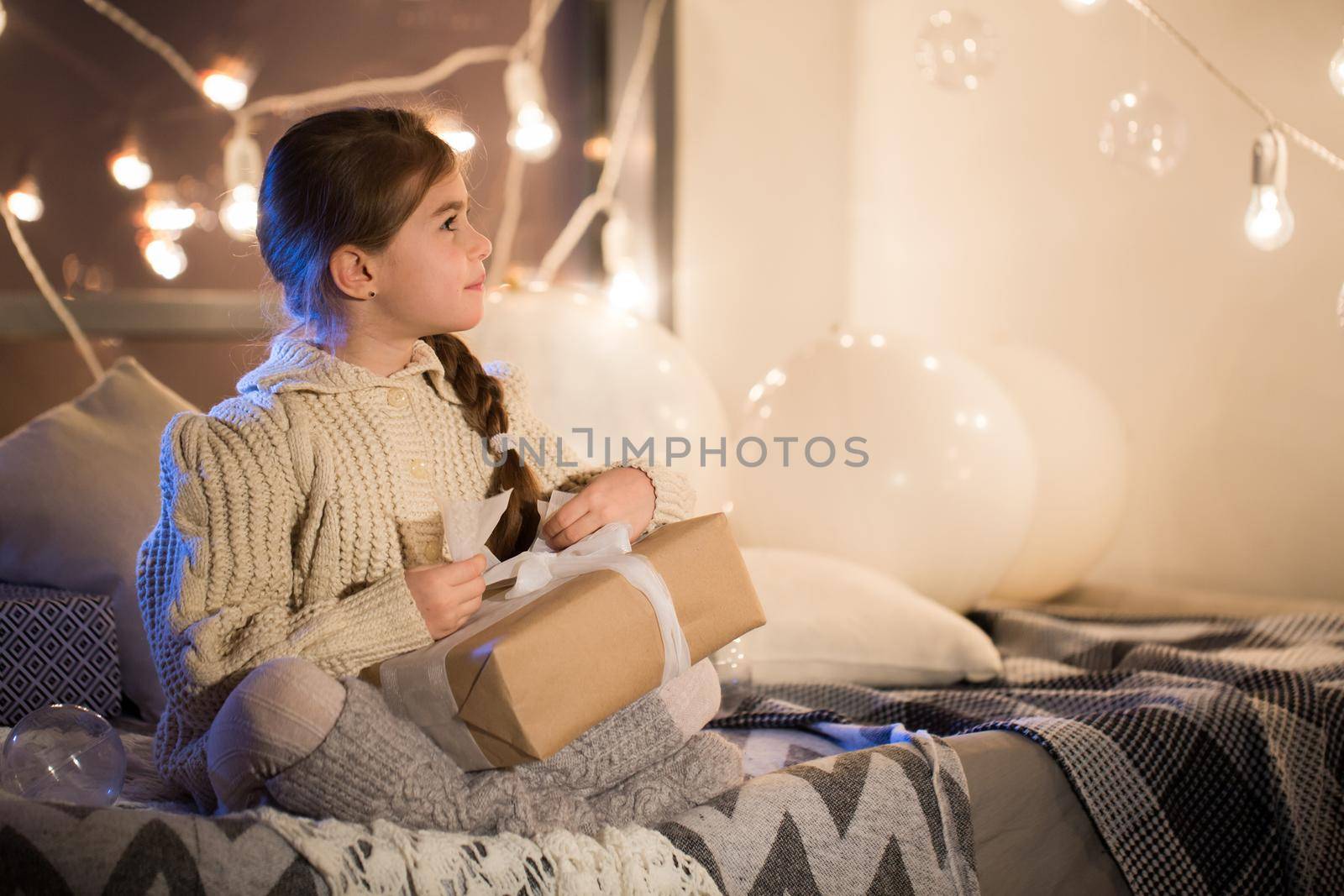 Beautiful little girl sitting on bed with a gift in its hands. Deploy Christmas gift box on holiday in the evening in the beautiful interior room. New year.