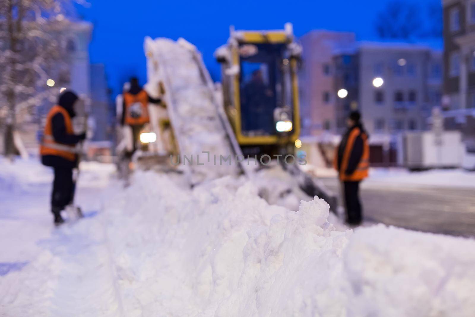 Claw loader vehicle removes snow from the road. Uniformed worker helps shovel snow into a snowplow by StudioPeace