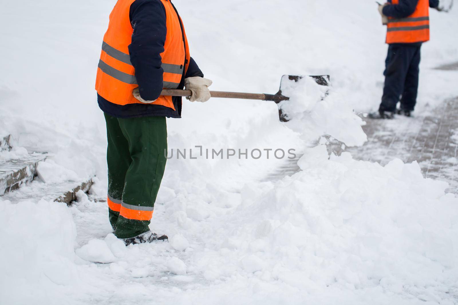 Employees of municipal services in a special form are clearing snow from the sidewalk with a shovel.