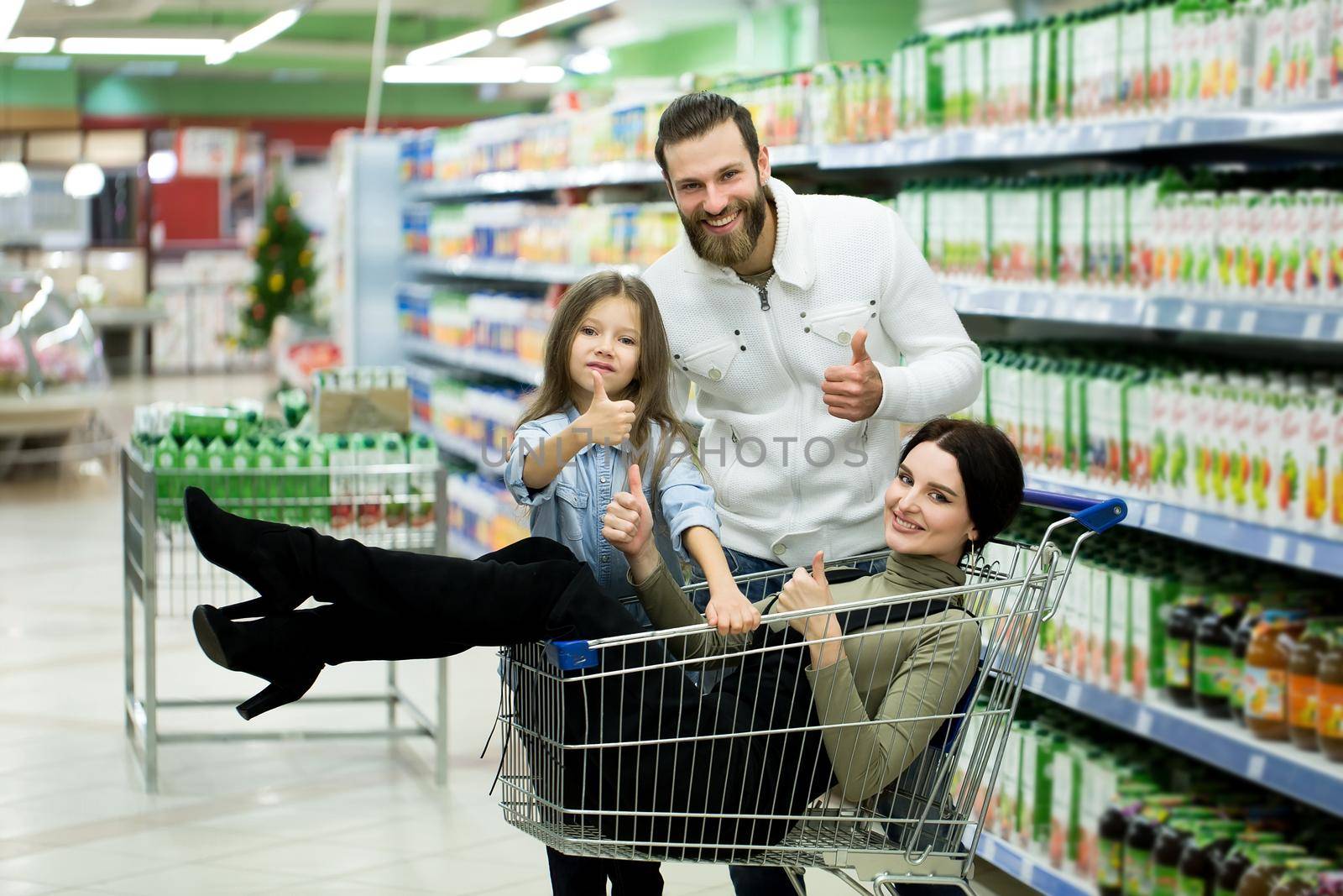 Beautiful young parents and their cute little daughter are smiling while choosing food in the supermarket. Father pushes the shopping cart while the girls sit there
