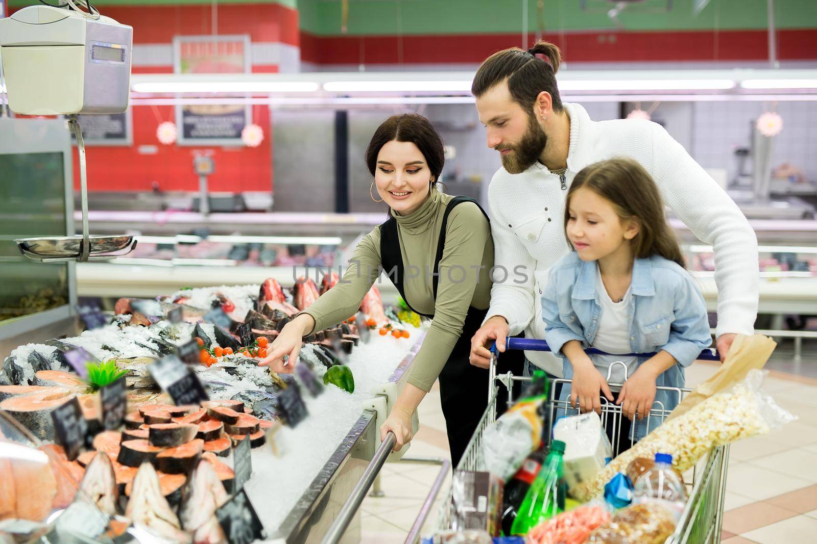 Beautiful young parents and their cute little daughter are smiling while choosing fish in the supermarket